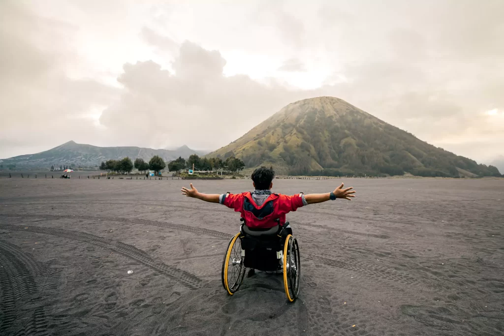ผู้พิการนั่งวีลแชร์มีฉากหลังเป็นทิวทัศน์อันตระการตาของปล่องภูเขาไฟที่พ่นควันสีขาวโพลน ท่ามกลางทะเลทรายทรายสีเทาและทิวเขาที่เรียงราย